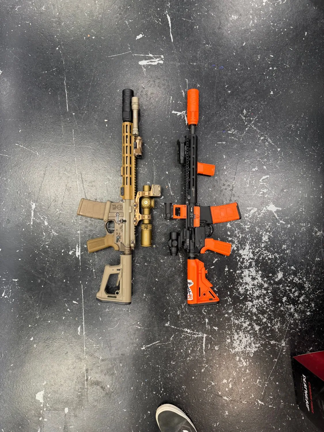 Two AR-style rifles side by side, one bronze and tan, one black with orange accents